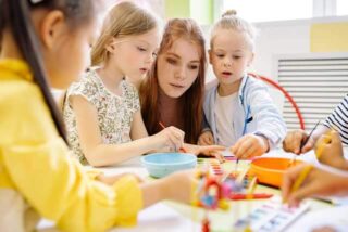 Two children using paintbrushes with their teacher during an art class with the help from a teacher